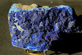 A raw blue lapis lazuli crystal specimen with deep indigo and cobalt blue matrix, showing natural texture and a light tan matrix area on top, displayed on a small stand against a dark background.