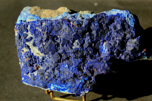 A raw blue lapis lazuli crystal specimen with deep indigo and cobalt blue matrix, showing natural texture and a light tan matrix area on top, displayed on a small stand against a dark background.