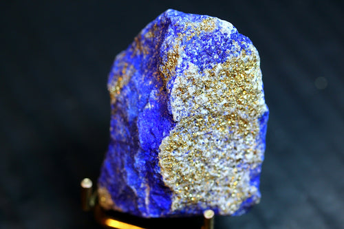 A raw blue lapis lazuli crystal specimen with prominent metallic gold pyrite inclusions, mounted on a small gold-colored 