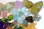 A collection of raw, rough-cut gemstones and crystals in various colors, including light blue, green, purple, pink, yellow, and clear quartz, arranged on a plain white background.