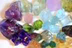 A close-up arrangement of assorted rough, faceted gemstones and crystals in multiple colors, including purple, blue, green, pink, yellow, and clear variants
