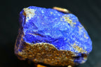 A close-up of a rough blue lapis stone or mineral specimen with metallic gold glittering along the lower edge and scattered patches