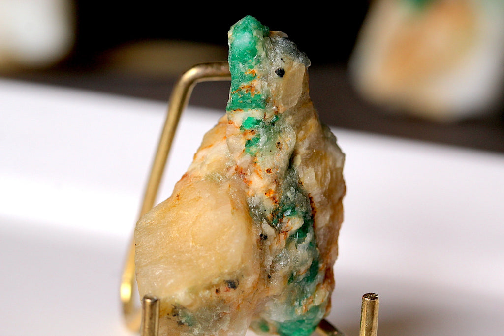 A raw mineral specimen with green and cream-colored matrix, displayed on a small gold-colored metal stand against a blurred white and black background.