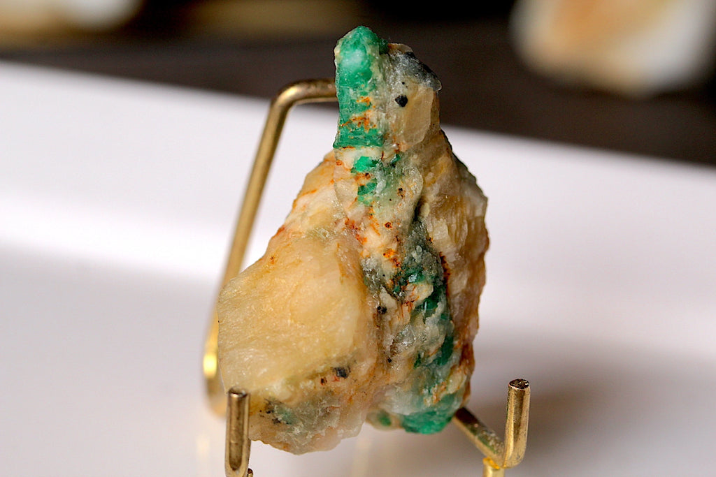A rough, natural gemstone specimen with a mix of pale cream, green, and orange-brown tones, displayed on a small gold-colored metal stand against a blurred white and dark background.