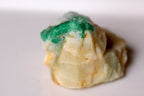 A raw, rough gemstone or mineral specimen with a vibrant green emerald from Swat top layer and a pale cream-colored body, resting on a plain white surface.
