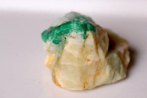 A raw, rough gemstone or mineral specimen with a vibrant green emerald from Swat top layer and a pale cream-colored body, resting on a plain white surface.