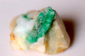 A raw green emerald stone or mineral specimen with a mix of vivid emerald green and milky white areas, resting on a plain light background.