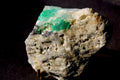 A raw emerald mineral specimen featuring vibrant green and pale green crystalline formations set in a rough, textured beige and gray matrix, photographed against a dark background.
