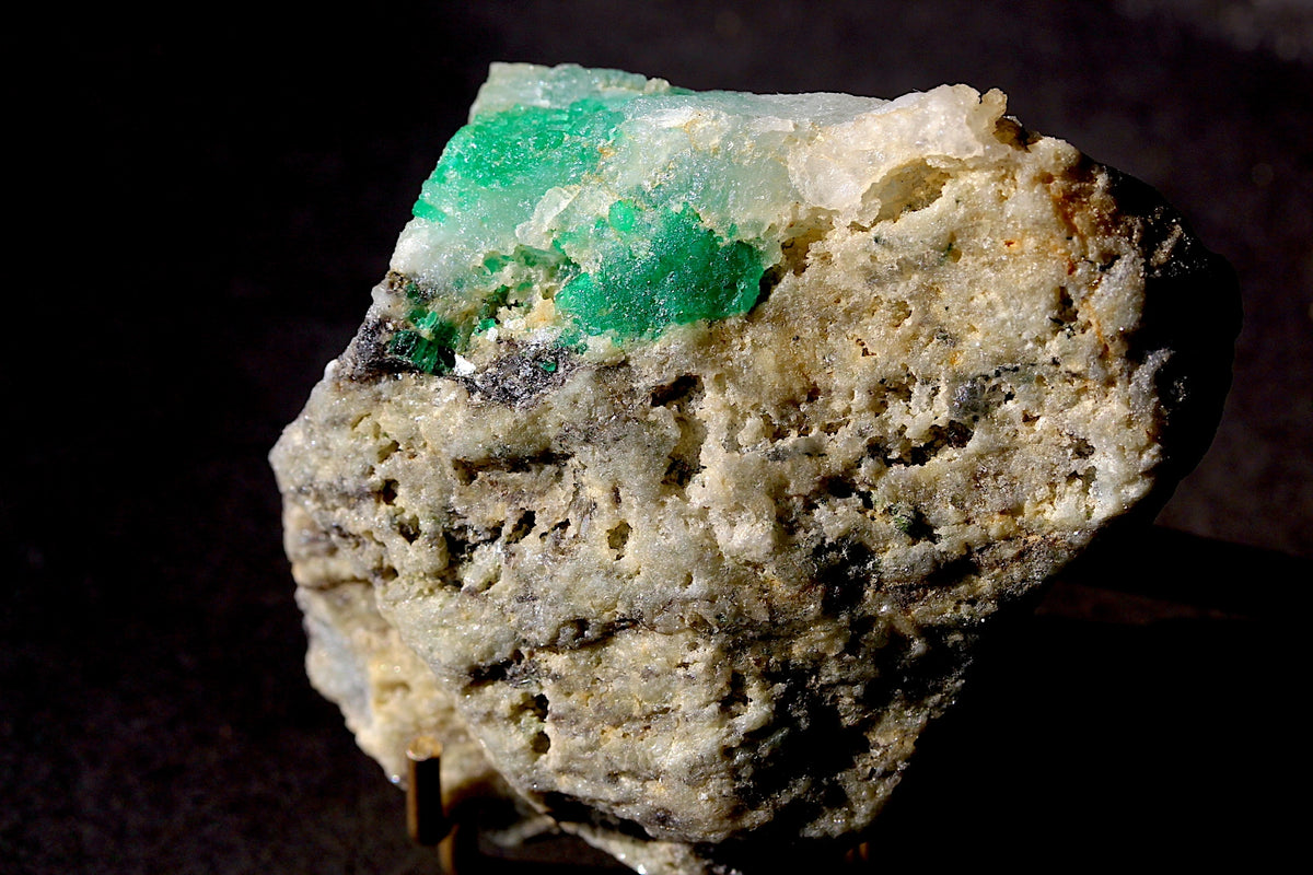A raw emerald mineral specimen featuring vibrant green and pale green crystalline formations set in a rough, textured beige and gray matrix, photographed against a dark background.