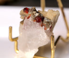 A raw crystal specimen with translucent white quartz and small red gemstone inclusions, displayed on a gold-colored metal stand against a white background.