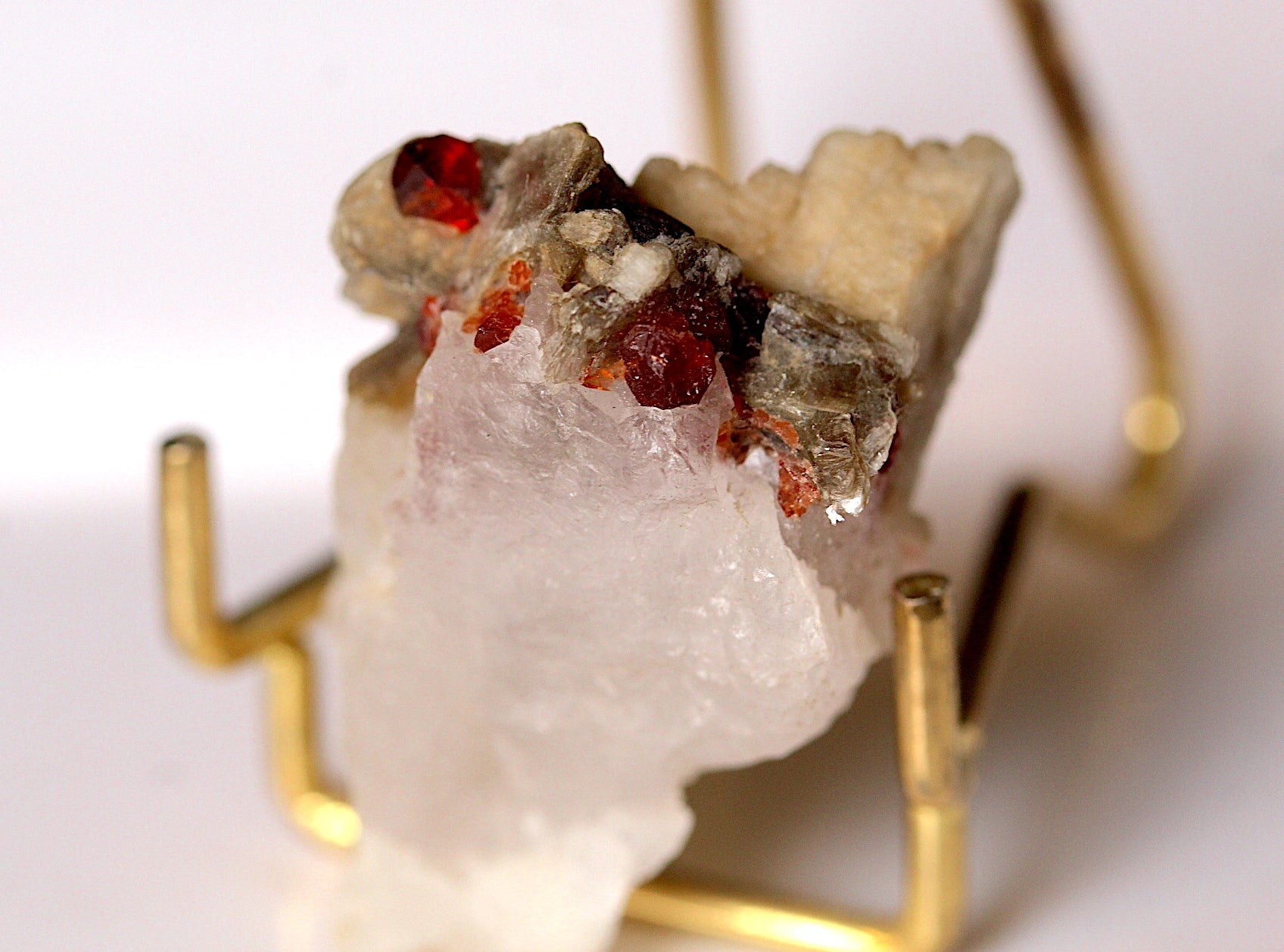 A raw mineral specimen featuring a large translucent white crystal section with rough, earthy gray-brown matrix and small red gemstone inclusions, displayed on a gold-colored metal stand against a white background.