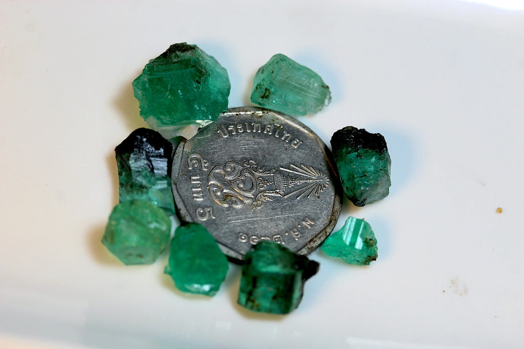 Several rough green emerald crystals arranged around a silver coin on a plain white surface.