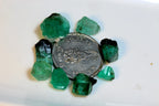 Several rough green emerald crystals arranged around a silver coin on a plain white surface.